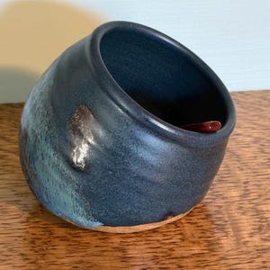May include: A handmade ceramic salt cellar with a dark blue glaze and a small wooden spoon. The bowl has a unique shape and a textured surface, with hints of turquoise and brown. The salt cellar is sitting on a wooden surface.