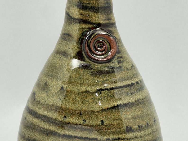 May include: A handcrafted ceramic vase with a unique, organic shape. The vase features a blend of olive green and dark brown horizontal stripes. A spiral design in shades of brown adorns the side, adding a decorative touch. The vase is a decorative item.