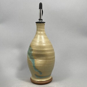 May include: A ceramic olive oil bottle with a light brown glaze and a blue swirl design. The bottle has a metal pour spout and a wooden base.
