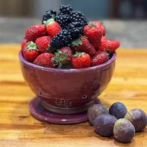May include: A purple ceramic colander bowl filled with fresh strawberries and blackberries. The bowl is sitting on a wooden surface with four ripe figs beside it.