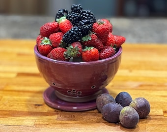 May include: A purple ceramic colander bowl filled with fresh strawberries and blackberries. The bowl is sitting on a wooden surface with four ripe figs beside it.