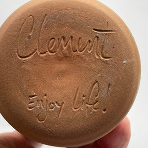 May include: A terracotta-colored ceramic pot with the words "Clement" and "Enjoy life!" inscribed on the bottom.