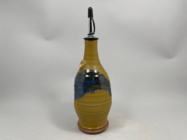 May include: A ceramic olive oil bottle with a brown and blue glaze. The bottle has a spout with a black and silver pourer.