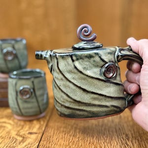 May include: Handmade ceramic tea set featuring a teapot with a spiral finial and handle, and two matching cups. The teapot and cups are glazed in shades of green and brown with a spiral design. The teapot is being held by a hand.