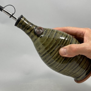 May include: A ceramic olive oil dispenser with a stainless steel spout and a black rubber stopper. The bottle has a green and brown striped pattern and a decorative swirl. The dispenser is held in a hand.