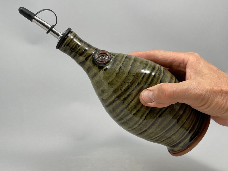 May include: A ceramic olive oil dispenser with a stainless steel spout and a black rubber stopper. The bottle has a green and brown striped pattern and a decorative swirl. The dispenser is held in a hand.