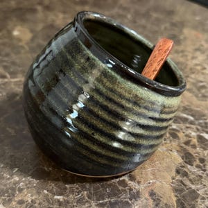 May include: A ceramic salt cellar with a wooden spoon. The small, round container has a dark green and black glazed finish with horizontal stripes. The spoon is a light brown color.