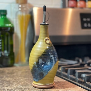 May include: A ceramic olive oil bottle with a black pour spout and a yellow and blue glaze. The bottle is sitting on a counter top in front of a stove.