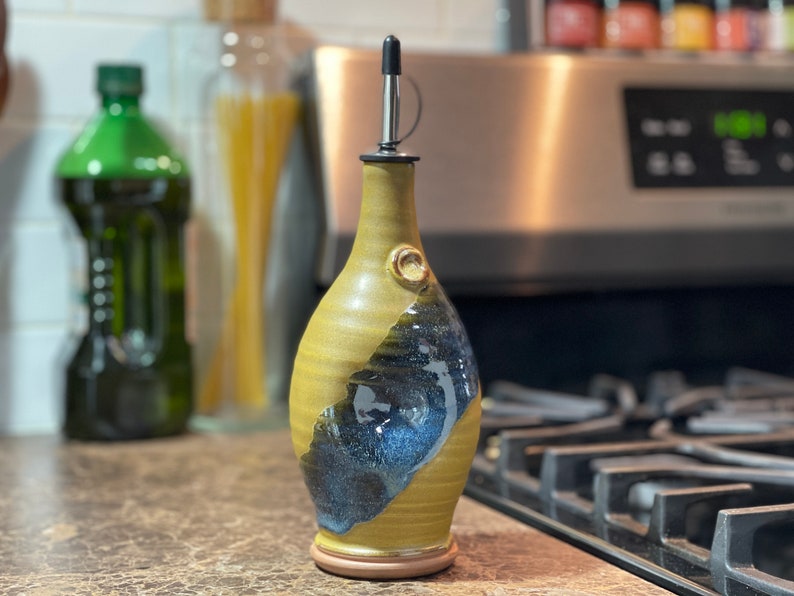 May include: A ceramic olive oil bottle with a black pour spout and a yellow and blue glaze. The bottle is sitting on a counter top in front of a stove.