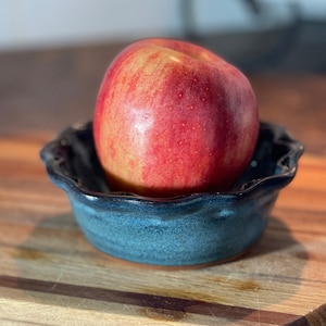May include: A red apple sits in a blue and white ceramic bowl with a scalloped rim. The bowl is on a wooden surface.
