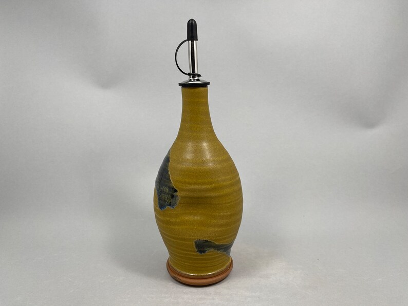 May include: A yellow ceramic oil and vinegar bottle with a black and blue glaze. The bottle has a stainless steel pour spout with a black rubber stopper.