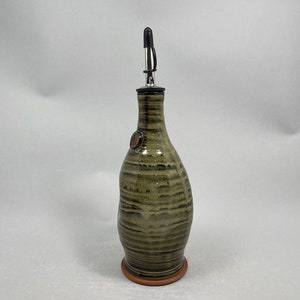 May include: A handcrafted olive oil bottle with a ceramic body and a stainless steel spout. The bottle has a green and brown striped pattern and a small, round emblem. It sits on a terracotta-colored base.