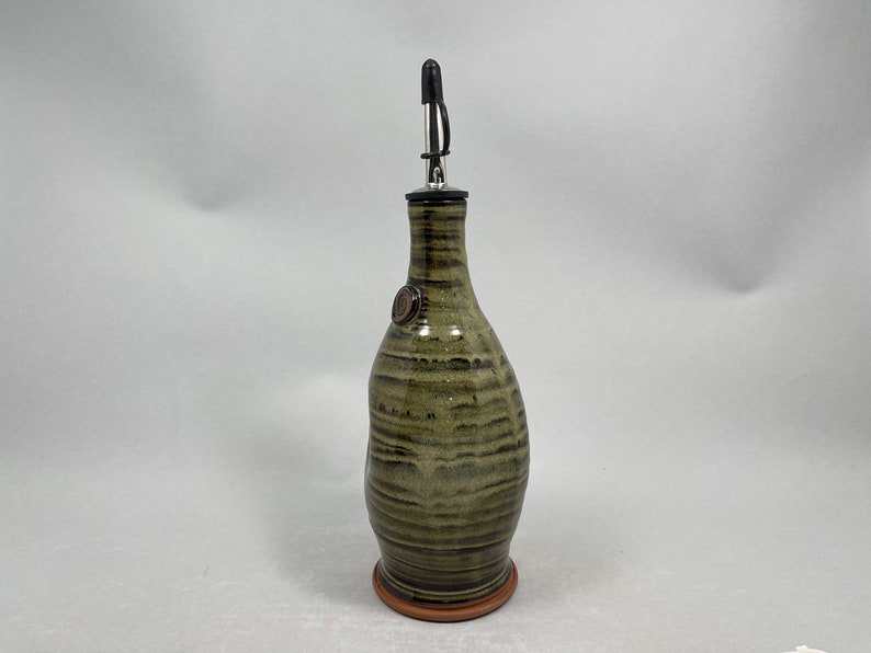 May include: A handcrafted olive oil bottle with a ceramic body and a stainless steel spout. The bottle has a green and brown striped pattern and a small, round emblem. It sits on a terracotta-colored base.
