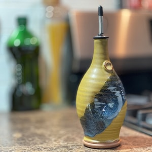 May include: A handmade ceramic olive oil bottle with a yellow and blue glaze. The bottle has a spout with a metal pourer.