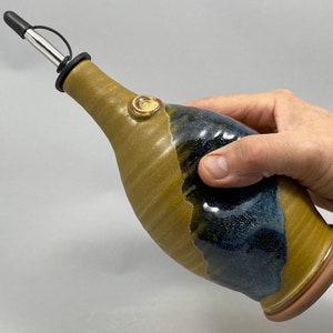 May include: A handmade ceramic olive oil bottle with a brown and blue glaze. The bottle has a spout with a black silicone tip and a metal pourer.