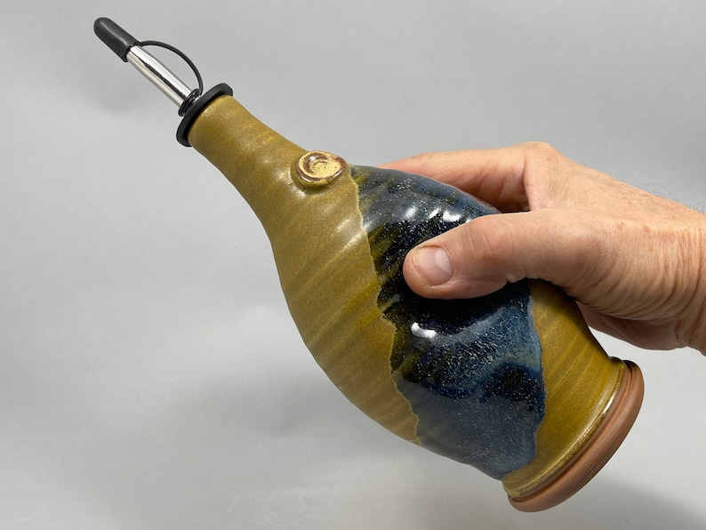 May include: A handmade ceramic olive oil bottle with a brown and blue glaze. The bottle has a spout with a black silicone tip and a metal pourer.