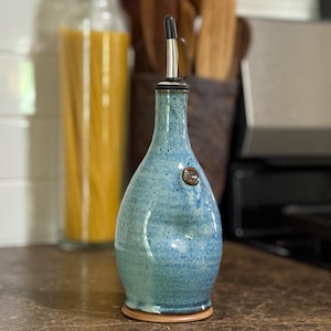 May include: A handmade, blue ceramic olive oil dispenser with a stainless steel spout. The bottle has a textured surface and a small, round, brown accent. The dispenser is on a brown base, and is in a kitchen setting.