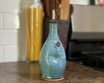 May include: A handmade, blue ceramic olive oil dispenser with a stainless steel spout. The bottle has a textured surface and a small, round, brown accent. The dispenser is on a brown base, and is in a kitchen setting.