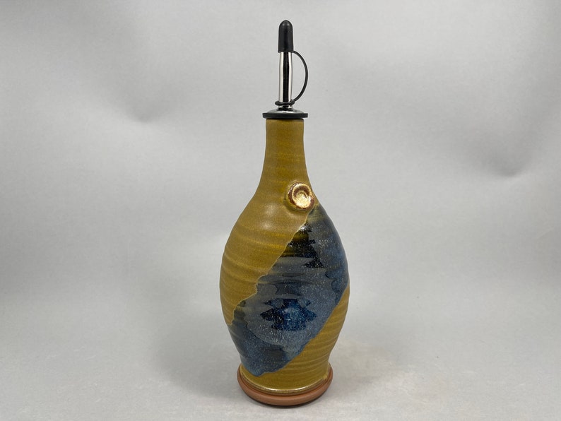 May include: A handmade ceramic olive oil bottle with a blue and yellow glaze. The bottle has a spout with a black stopper and a small knob on the side.