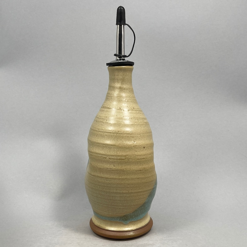 May include: A ceramic olive oil bottle with a light brown and blue glaze. The bottle has a metal pour spout with a black stopper.