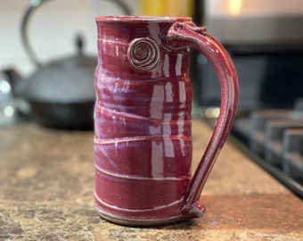 May include: A handmade ceramic mug with a burgundy glaze and a swirl design on the handle. The mug has a unique, textured surface.