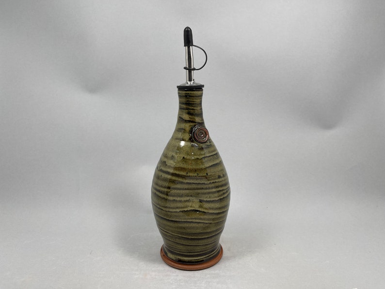 May include: A handcrafted ceramic olive oil bottle with a dark green and brown striped pattern. It features a stainless steel spout with a black cap and a decorative spiral accent. The bottle stands approximately 10 inches tall.
