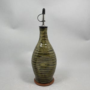 May include: A ceramic olive oil bottle with a dark metal spout and a brown base. The bottle has a textured, earthy green and brown striped design. The spout has a black cap.