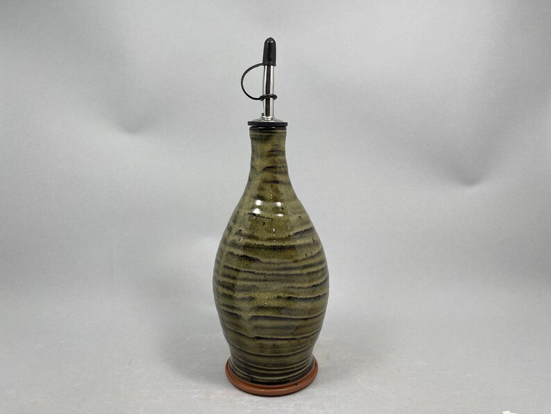 May include: A ceramic olive oil bottle with a dark metal spout and a brown base. The bottle has a textured, earthy green and brown striped design. The spout has a black cap.