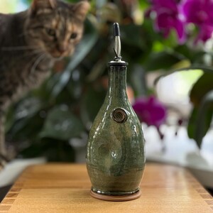 May include: A green and brown ceramic oil bottle with a metal spout and a decorative swirl. The bottle sits on a wooden surface, with a cat and purple flowers in the background.