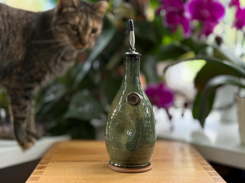 May include: A green and brown ceramic oil bottle with a metal spout and a decorative swirl. The bottle sits on a wooden surface, with a cat and purple flowers in the background.