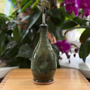 May include: A green and brown ceramic oil bottle with a stainless steel spout. The bottle has a rounded shape and sits on a wooden base. The background features green plants and purple flowers.