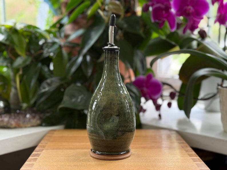 May include: A green and brown ceramic oil bottle with a stainless steel spout. The bottle has a rounded shape and sits on a wooden base. The background features green plants and purple flowers.