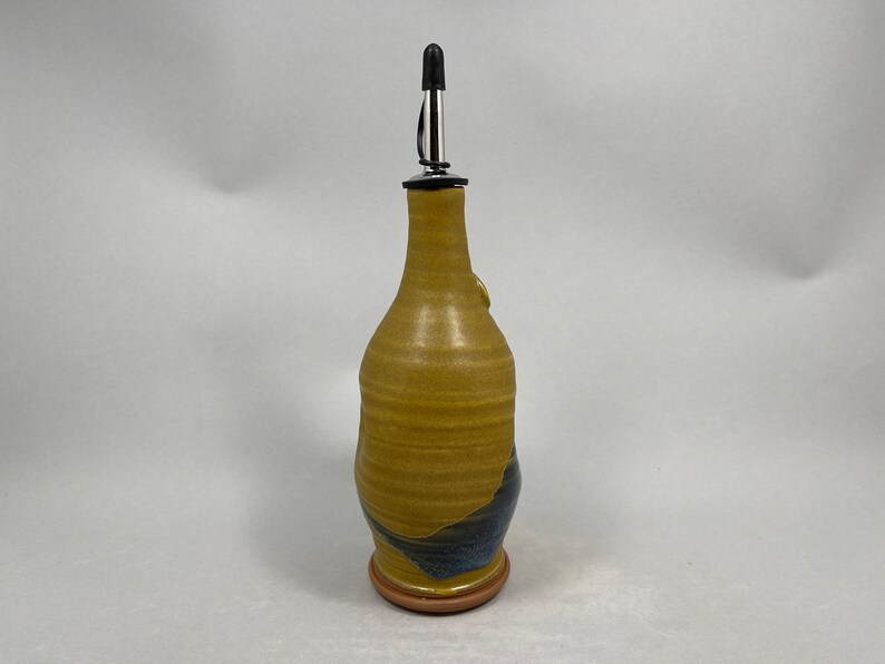 May include: A ceramic olive oil bottle with a yellow and blue glaze. The bottle has a stainless steel pour spout with a black rubber stopper.