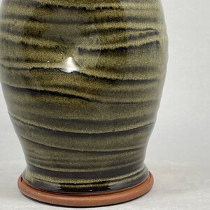 May include: A handmade ceramic vase with a dark olive green and black striped pattern. The vase has a wide body that tapers towards the base, which sits on a terracotta-colored base. The vase is glazed and has a glossy finish.