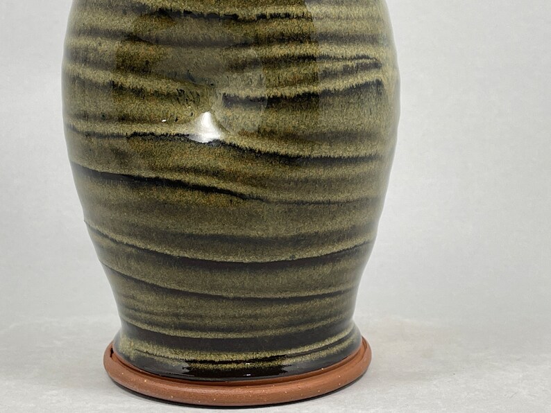 May include: A handmade ceramic vase with a dark olive green and black striped pattern. The vase has a wide body that tapers towards the base, which sits on a terracotta-colored base. The vase is glazed and has a glossy finish.
