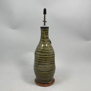 May include: A handcrafted ceramic olive oil bottle with a stainless steel spout. The bottle has a textured, earthy green and brown glaze with horizontal stripes. It sits on a small, round, terracotta-colored base.