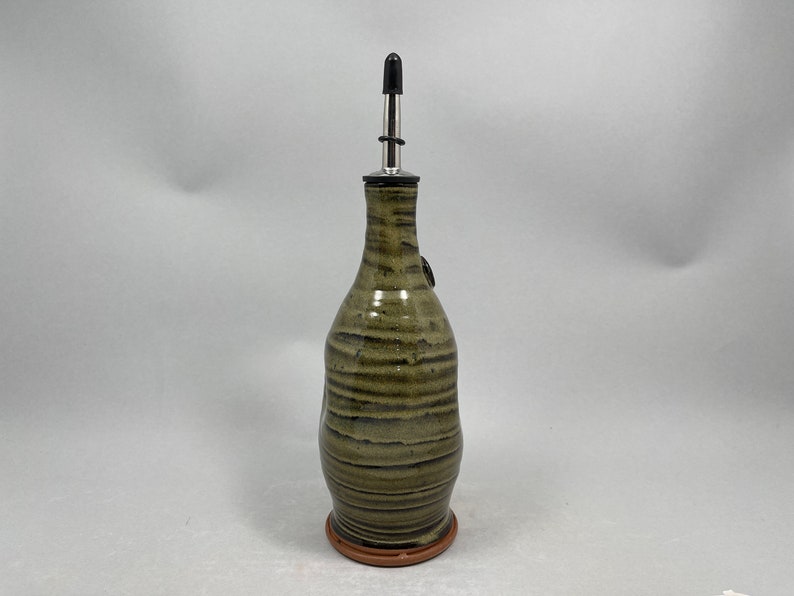 May include: A handcrafted ceramic olive oil bottle with a stainless steel spout. The bottle has a textured, earthy green and brown glaze with horizontal stripes. It sits on a small, round, terracotta-colored base.