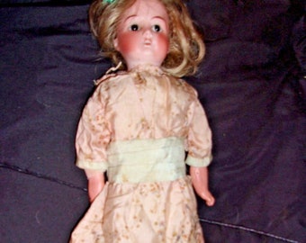 May include: A vintage porcelain doll with blonde hair, blue eyes, and a pink dress with floral print. The doll is wearing black shoes and a green ribbon in her hair.