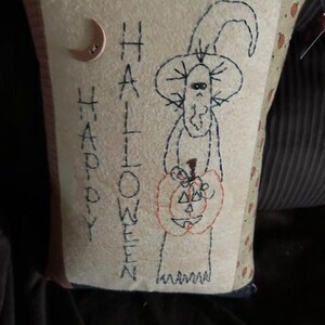 May include: A beige pillow with a stitched design of a witch holding a pumpkin. The text "HAPPY HALLOWEEN" is stitched on the pillow.