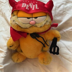 May include: A plush Garfield toy dressed as a devil. The orange cat wears a red devil hat with the words "Little Devil" and a matching cape. He holds a black pitchfork and has a grumpy expression.