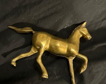 May include: A brass figurine of a horse in a running pose. The sculpture is a golden color and has detailed features, including a flowing tail and a defined mane. The horse is depicted with its legs extended, suggesting movement.