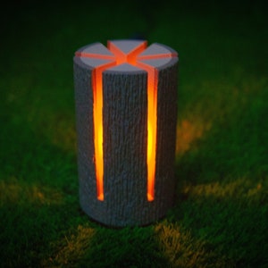 May include: A wooden lamp shaped like a log with a glowing orange light shining through the cutouts. The lamp is sitting on a bed of green grass.