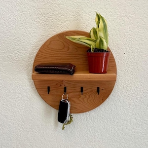 May include: A round wooden key holder with a shelf and five hooks. A brown leather wallet is on the shelf, and a small potted plant is on the right side of the shelf. A key is hanging from one of the hooks.