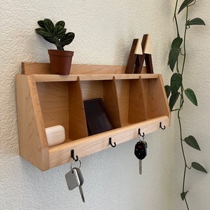 May include: A wooden wall-mounted organizer with four hooks and three compartments. The organizer is made of light wood and has a natural finish. There are keys, a wallet, and a small white box in the organizer.