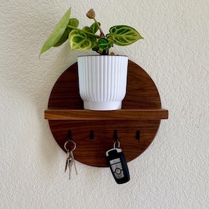 May include: A round wooden key holder with a shelf for a small plant. The key holder has four hooks and is mounted on a white wall. A house key and a car key are hanging on the hooks.