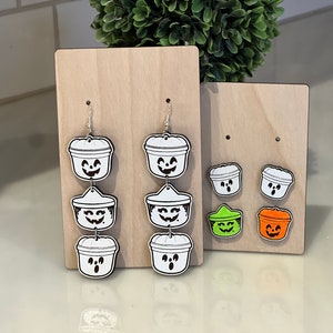May include: Halloween-themed earrings and stud earrings. The earrings are shaped like jack-o'-lantern buckets with smiling faces. The stud earrings are also jack-o'-lantern buckets, with green and orange options. All are displayed on wooden stands.