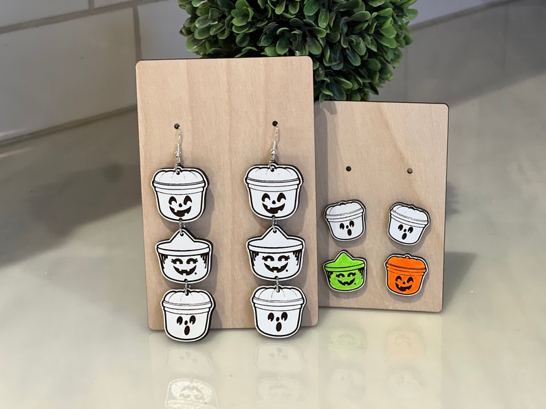 May include: Halloween-themed earrings and stud earrings. The earrings are shaped like jack-o'-lantern buckets with smiling faces. The stud earrings are also jack-o'-lantern buckets, with green and orange options. All are displayed on wooden stands.