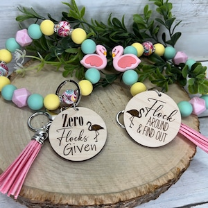 Puede incluir: Dos llaveros de flamenco rosa con etiquetas de madera. Una etiqueta dice "Zero Flocks Given" y la otra dice "Flock Around & Find Out". Los llaveros están unidos a una pulsera de cuentas de colores.