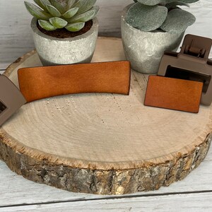May include: Two brown leather hair clips, one curved and one rectangular, on a wooden surface. The clips are next to two small potted plants.