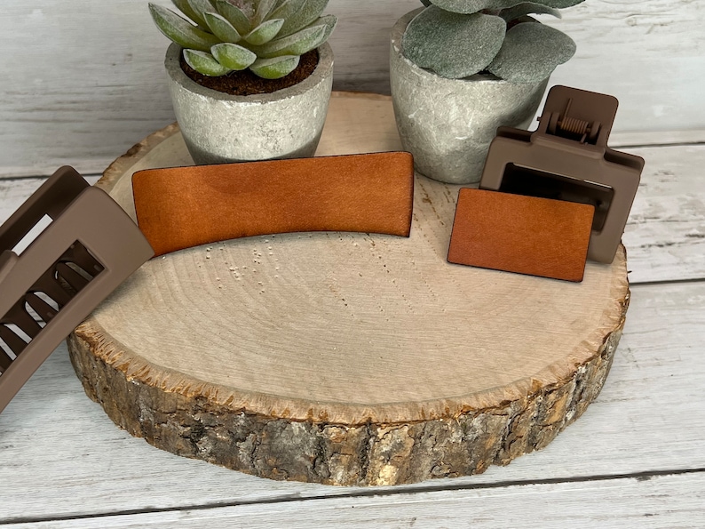 May include: Two brown leather hair clips, one curved and one rectangular, on a wooden surface. The clips are next to two small potted plants.